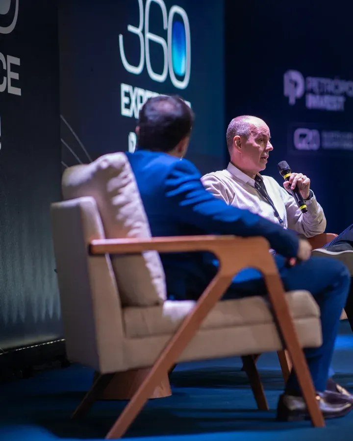 Dois convidados em poltronas no palco, um deles falando ao microfone