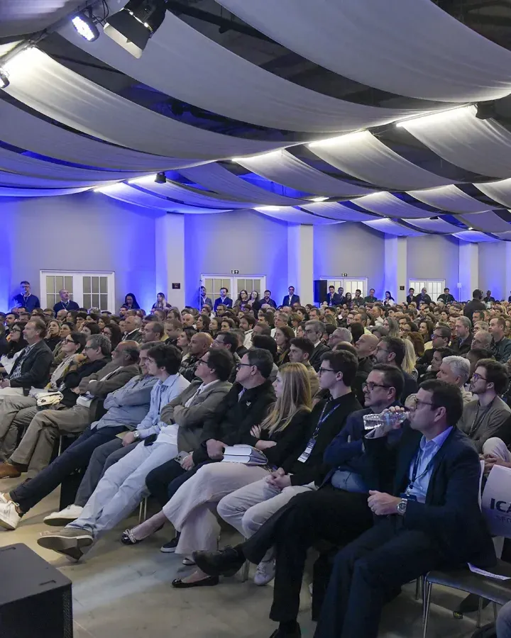 Público sentado nas cadeiras do auditório da 1ª edição, sob a luz azul do salão